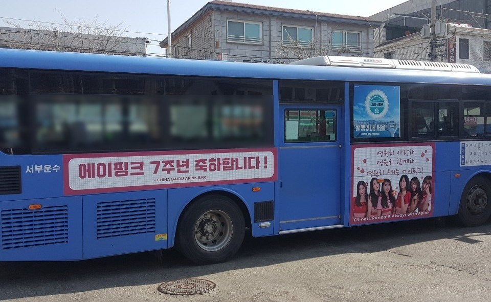 Door side of the same Seoul city bus with a wide pink banner reading "Congratulations on Apink's 7th anniversary" in Korean and a second panel with a group photo