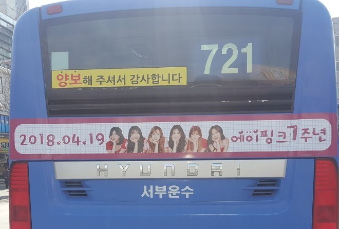 Rear of the bus with a strip ad showing the date 2018.04.19, a row of member photos and "Apink 7th anniversary" in Korean