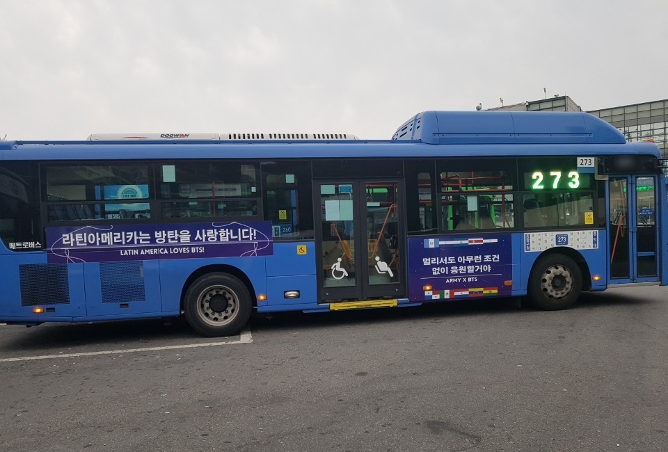 Door side of the bus with a purple banner reading "Latin America loves BTS!" in Korean and English and a second panel with small national flags