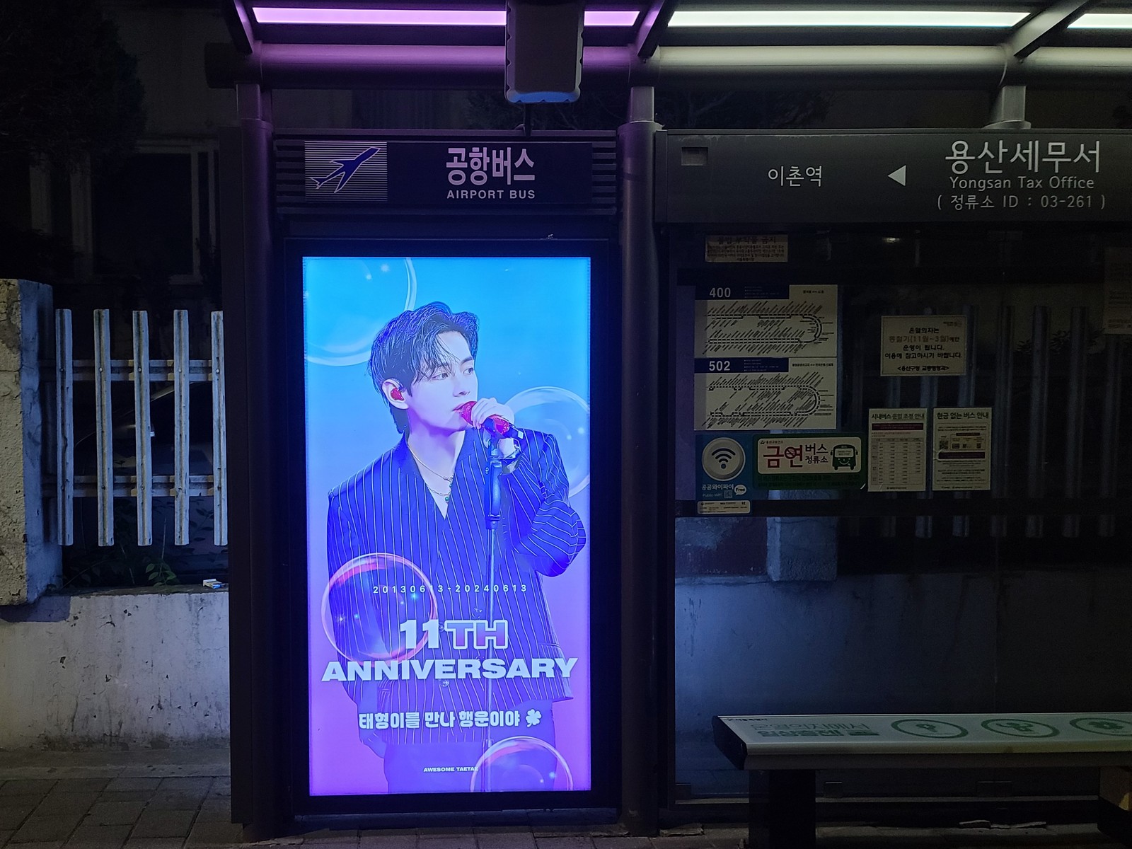 BTS V anniversary ad on an upright digital screen at a Seoul bus shelter at night, with a portrait at a microphone and the words 11TH ANNIVERSARY