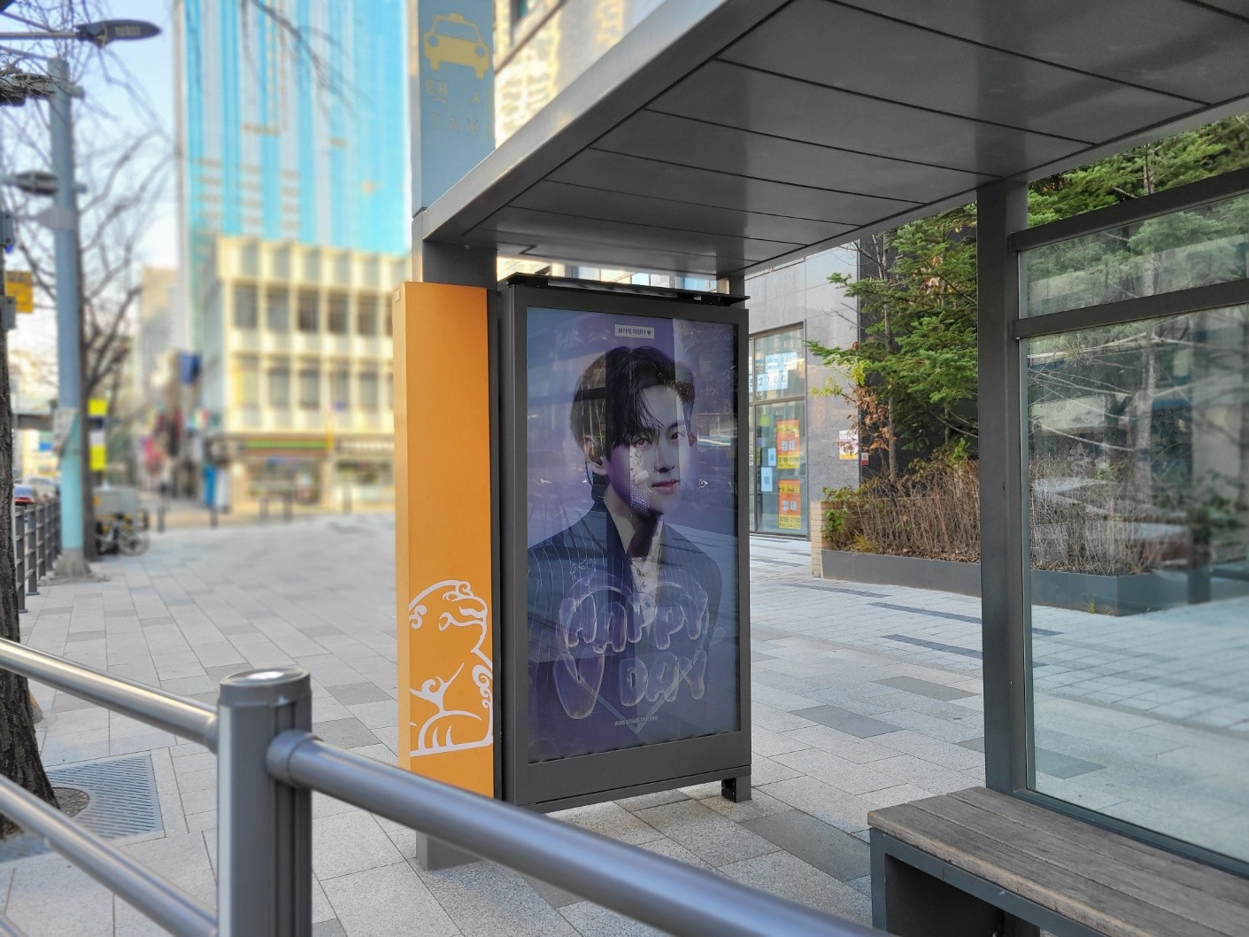 Wider view of a glass shelter with a bench and a taxi sign on the pole above, the screen fixed to its end wall