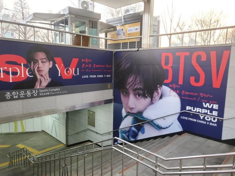 BTS V fan support ad wrapping two walls of a stairway entrance at Sports Complex Station in Seoul, with large red "BTS V" lettering on a navy background