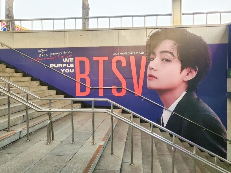 Sloping stairway wall covered by a navy print with red "BTS V" letters, a large portrait on the right and "We Purple You" in white
