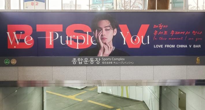 Banner above the station name sign reading "Sports Complex", with "BTS V" in red, "We Purple You" in white and "LOVE FROM CHINA V BAR"