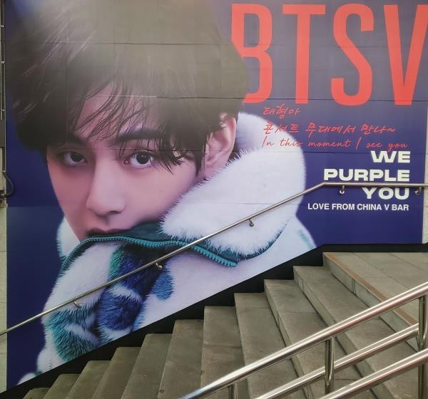 Close view of the facing wall with a portrait in a white fur collar, "BTS V" in red and "WE PURPLE YOU" beside the handrail