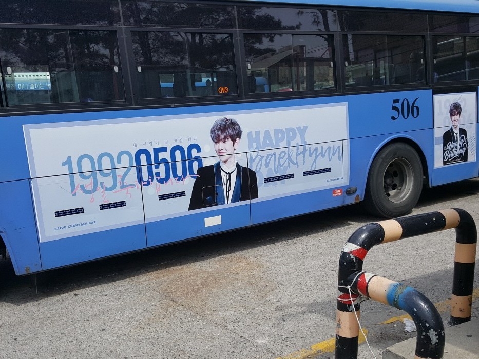 EXO Baekhyun birthday ad on the road side of a blue Seoul city bus, a pale banner with his photo, the numbers 19920506 and the words "Happy BaekHyun Day"