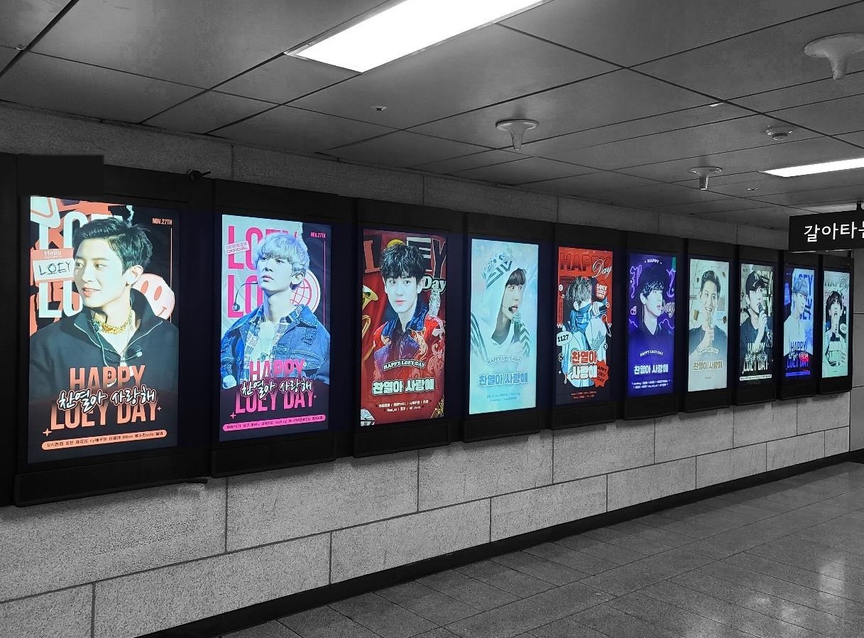 A row of ten upright digital screens along a station passage showing different "HAPPY LOEY DAY" designs