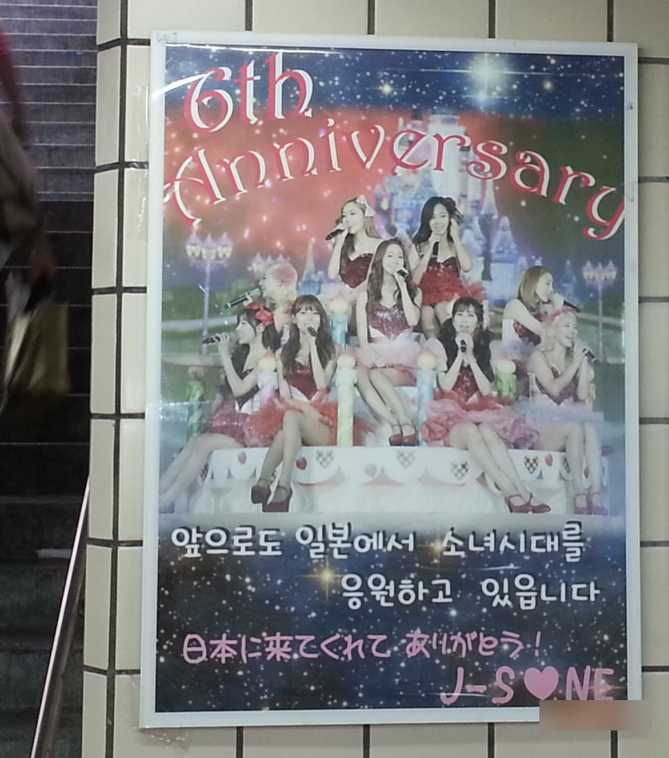 Girls' Generation 6th anniversary ad poster on a tiled subway station wall beside a staircase, with a group photo and a message in Korean and Japanese