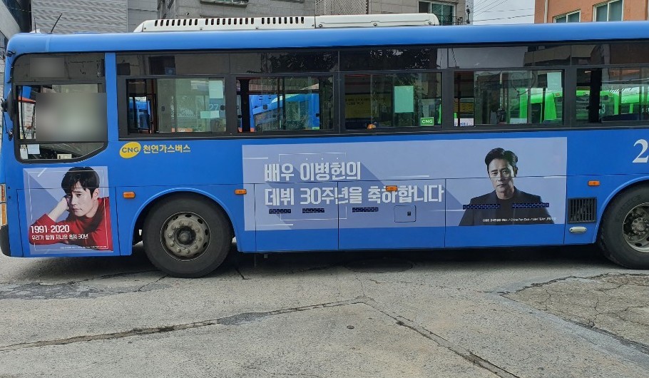 Lee Byung Hun debut anniversary ad on the side of a blue Seoul city bus, a pale blue banner with a Korean headline and a portrait in a dark suit
