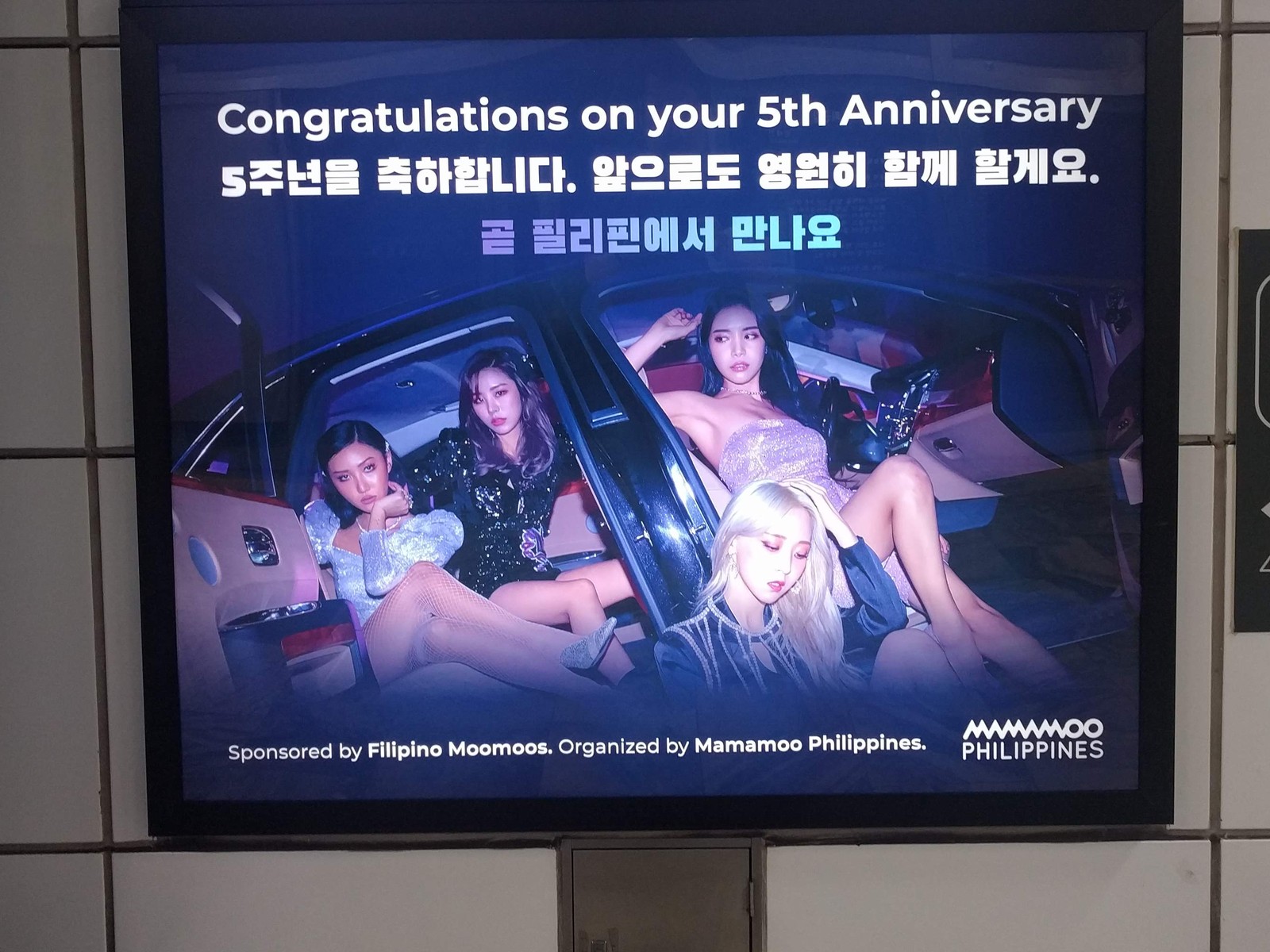 MAMAMOO anniversary ad on a framed screen in a Seoul subway station, with a group photo and the words Congratulations on your 5th Anniversary in English and Korean