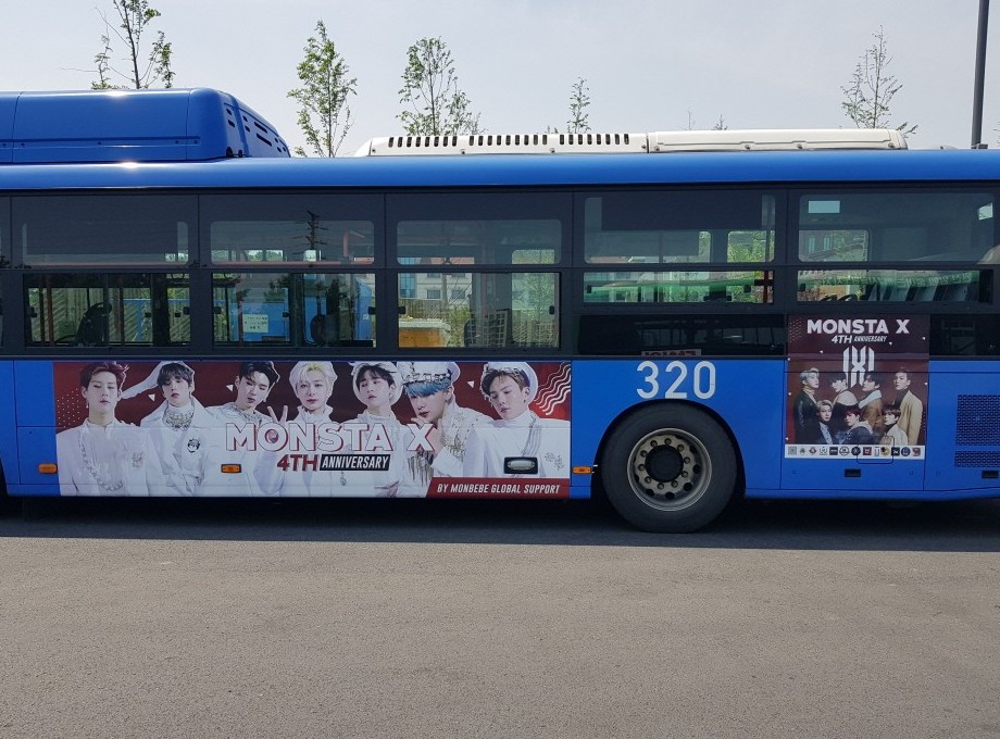 MONSTA X anniversary ad on the road side of a blue Seoul city bus, with seven members in white outfits and the words "MONSTA X 4TH ANNIVERSARY"