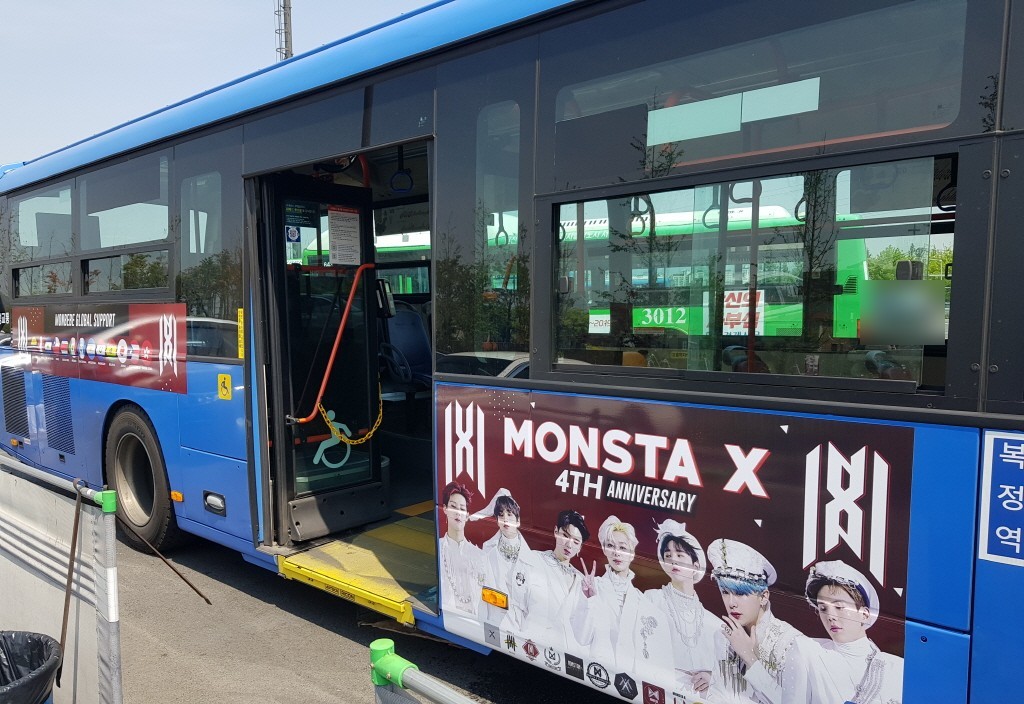 Door side of the same bus with a dark red panel reading "MONSTA X 4TH ANNIVERSARY" and a banner with a row of round flag badges