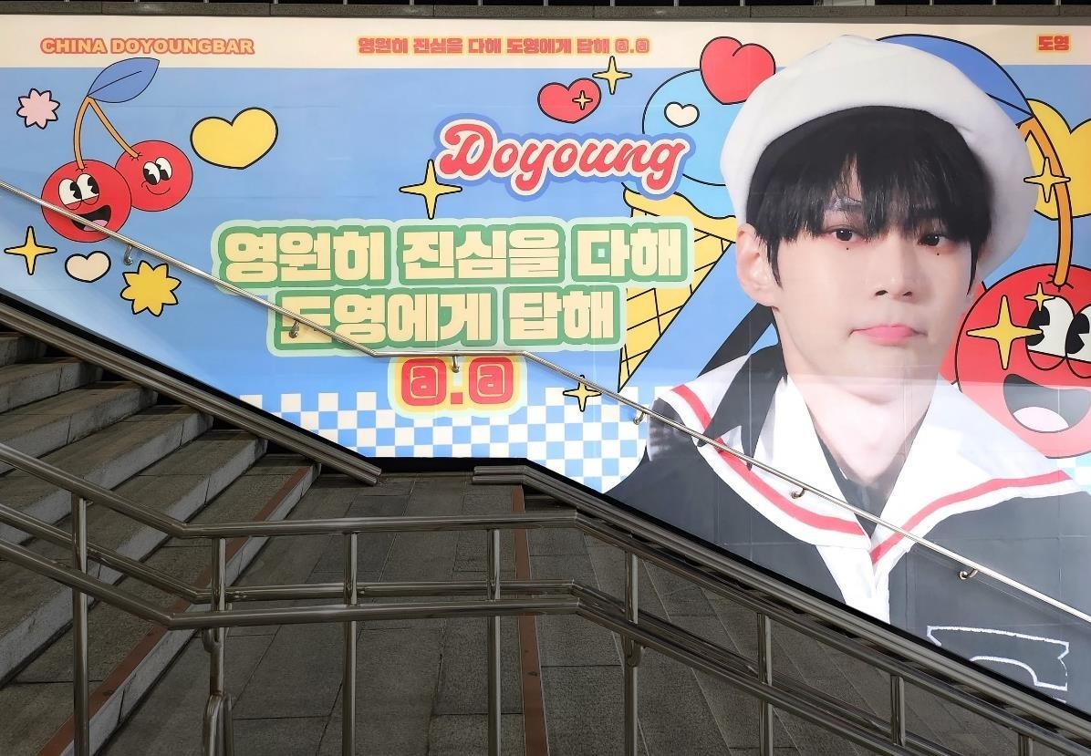 NCT Doyoung fan support ad on a stairway wall at Sports Complex Station in Seoul, with cartoon cherries and hearts, a green Korean message and a large photo of a singer in a white beret