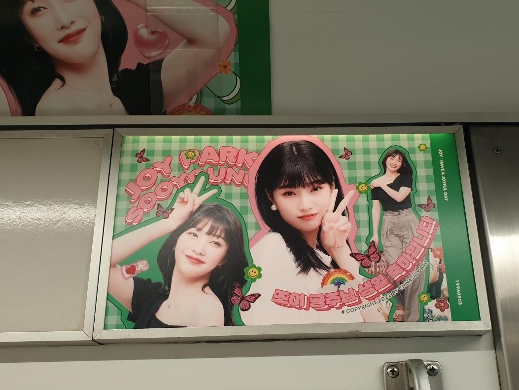 Joy birthday ad in a frame inside a subway car, a green checked poster with three photos and the name "JOY PARK SOOYOUNG"