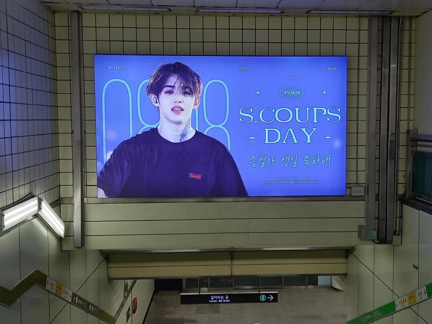 SEVENTEEN S.Coups birthday ad in a lit frame above a stairway in a Seoul subway station, with a stage photo on the left and "S.COUPS DAY" in pale green letters on violet blue